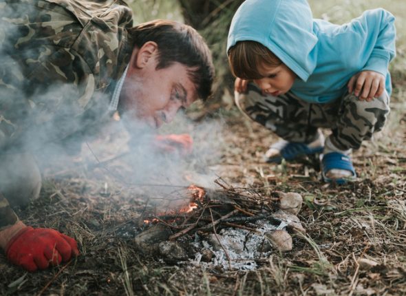 little happy five year old kid boy and his father blowing and kindles a campfire in the village or forest on nature outdoor on a summer evening. authentic countryside and children's rustic life