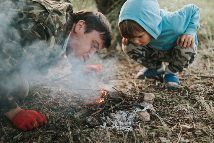 little happy five year old kid boy and his father blowing and kindles a campfire in the village or forest on nature outdoor on a summer evening. authentic countryside and children's rustic life